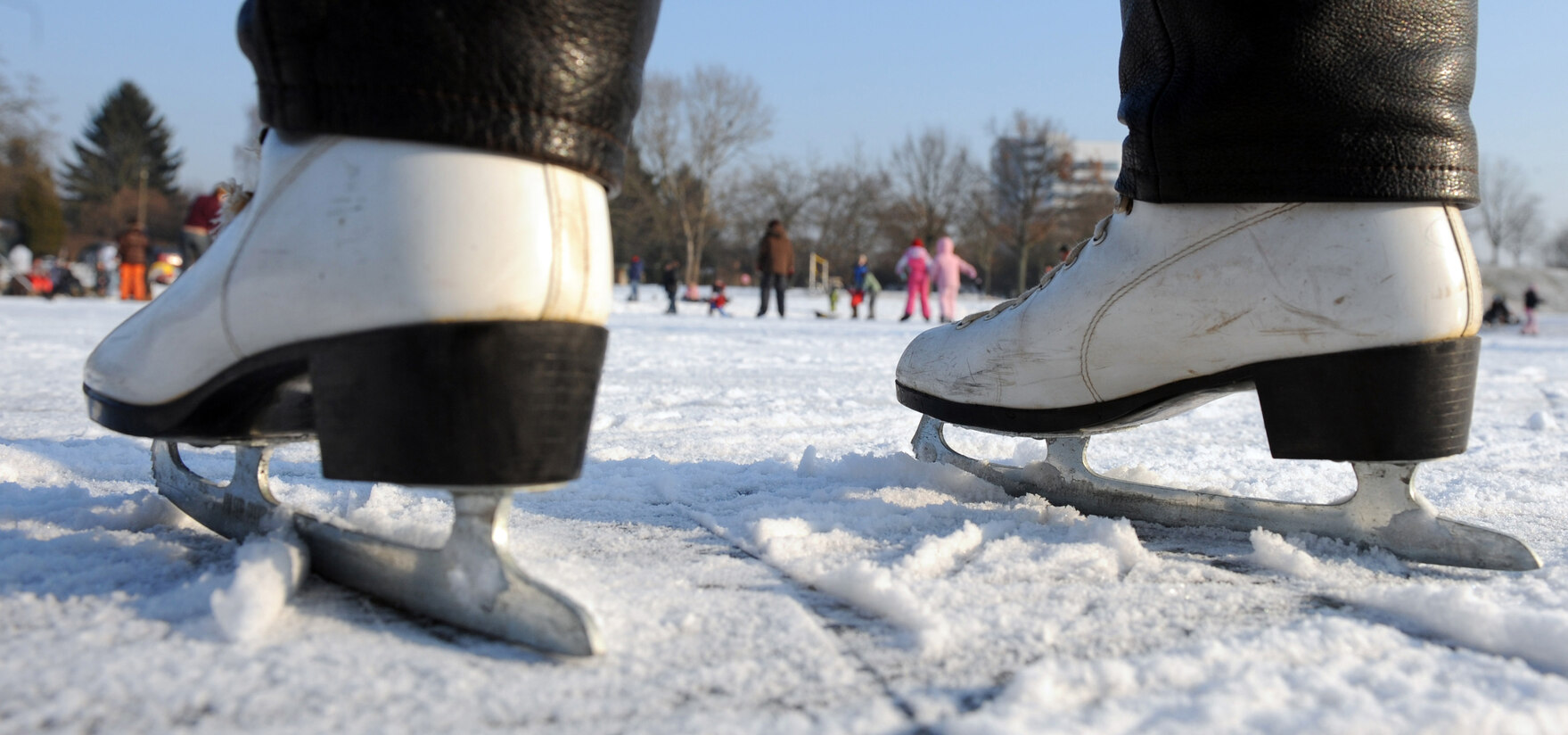 Ein Mensch mit Schlittschuhen steht auf einer Eisfläche, im Hintergrund sind Kinder zu sehen.